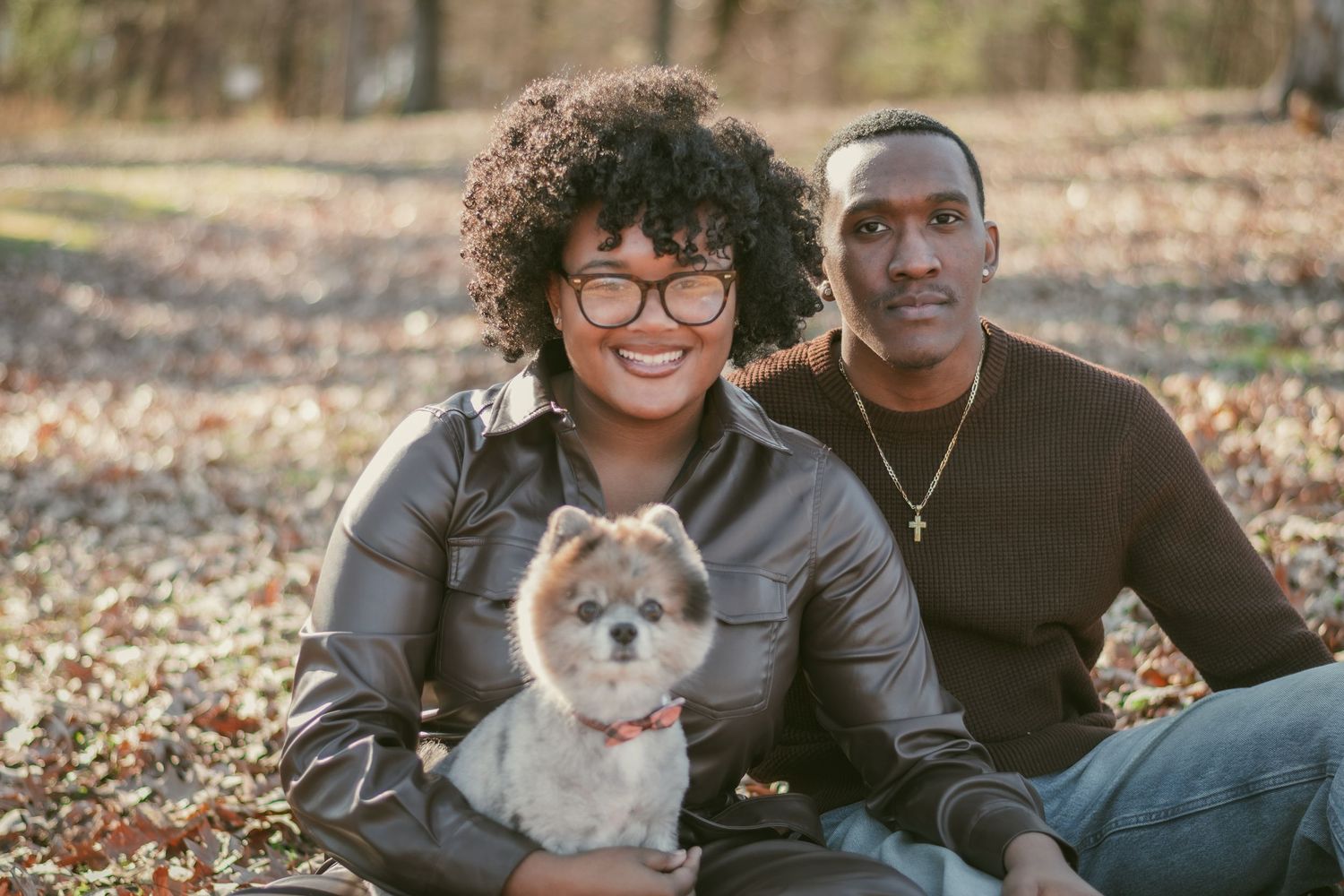Smiling couple sitting outdoors with a small Pomeranian dog, surrounded by autumn leaves in a park setting.