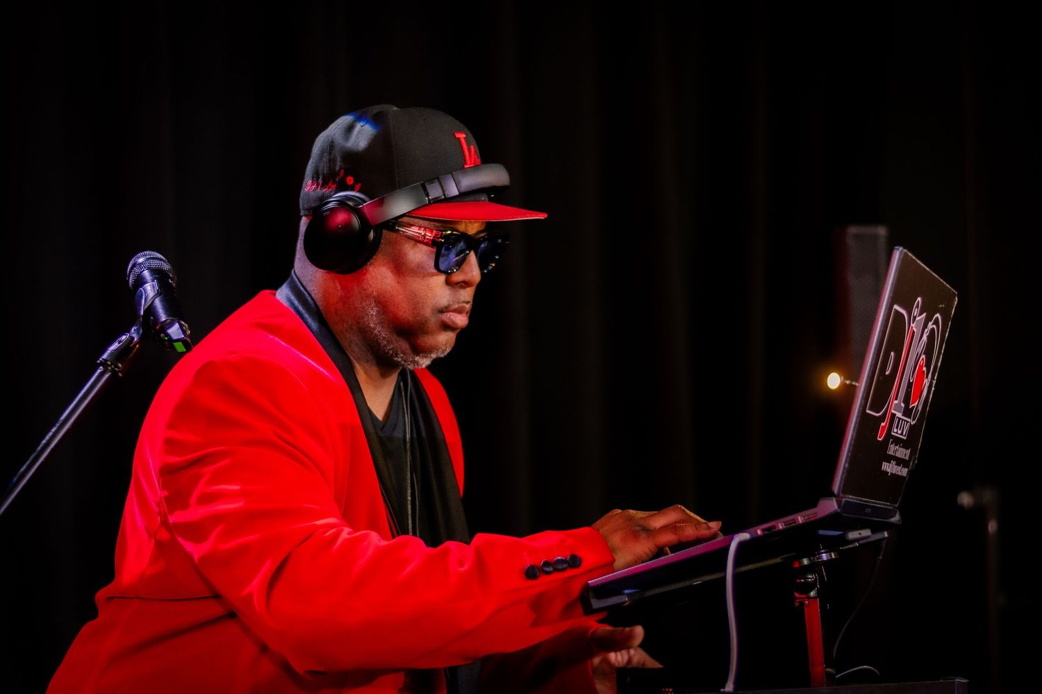 DJ in red jacket and headphones mixing music on a laptop at a live event performance.