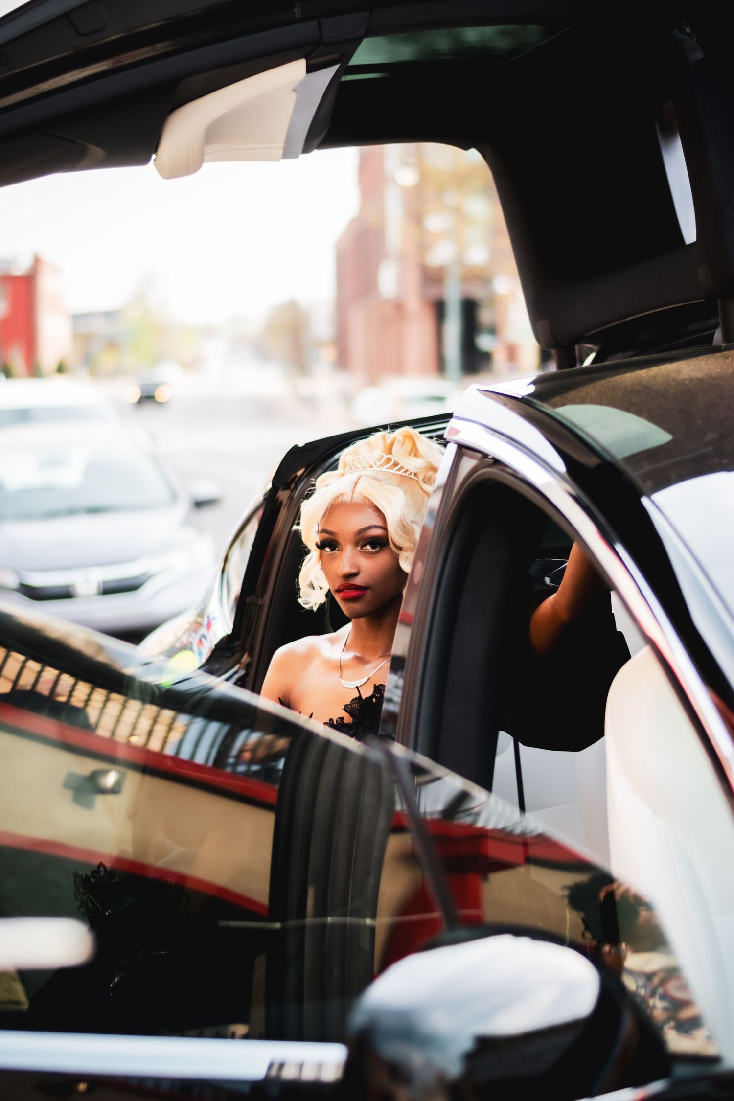 Elegant woman wearing a gold crown and jewelry, seen through a luxury car window in an urban setting.