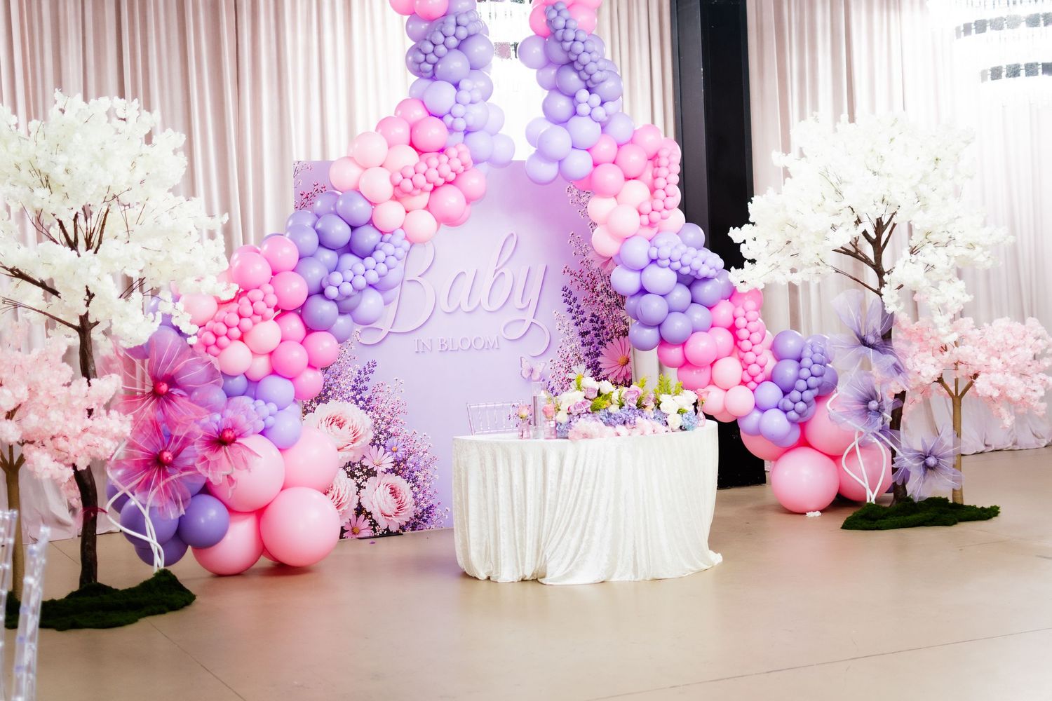 Pink and purple balloon arch frames a dessert table at a baby shower event.