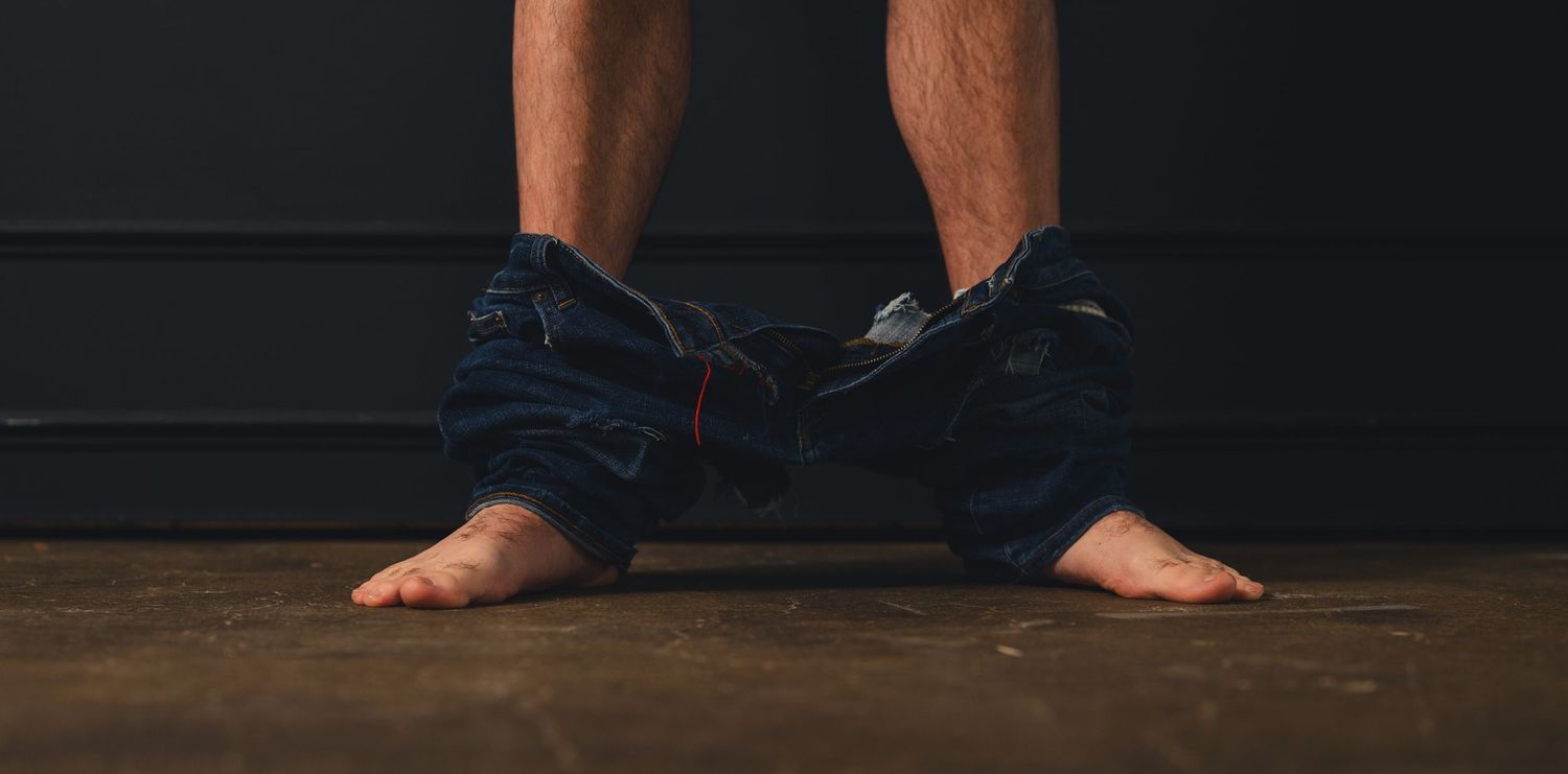 Person standing barefoot with jeans pulled down around ankles on cement floor against dark wall.