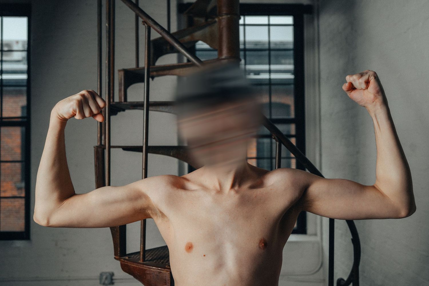Shirtless muscular man flexing biceps with blurred face near spiral staircase in industrial loft.