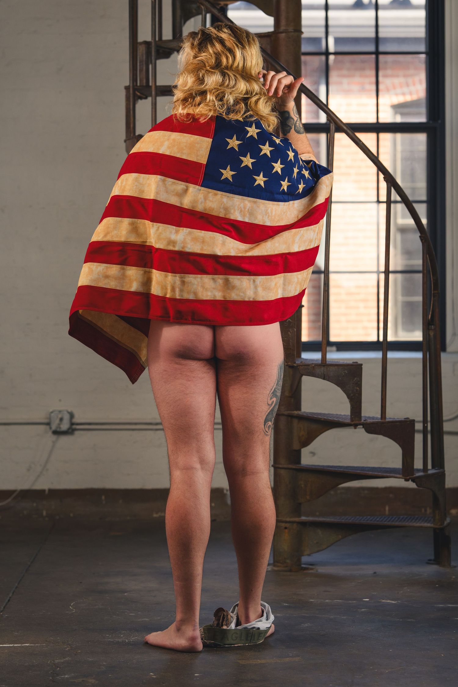 Man draped in vintage American flag standing barefoot near spiral staircase in industrial loft space.