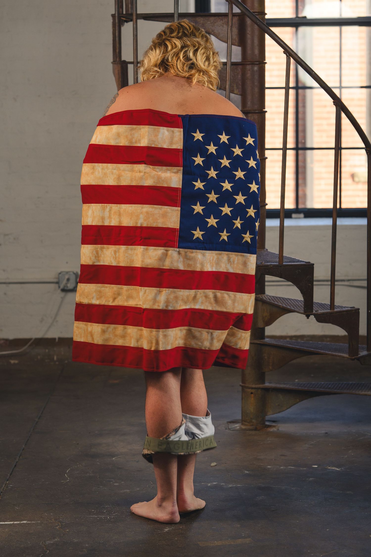 Person wrapped in an American flag standing barefoot near a spiral staircase indoors.