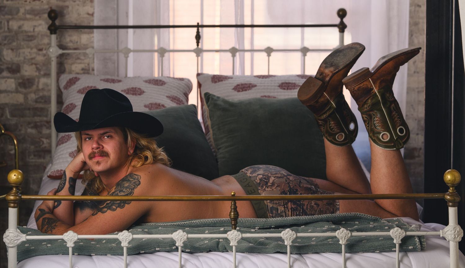 Tattooed man in black cowboy hat and boots lying on a vintage metal bed with decorative pillows.
