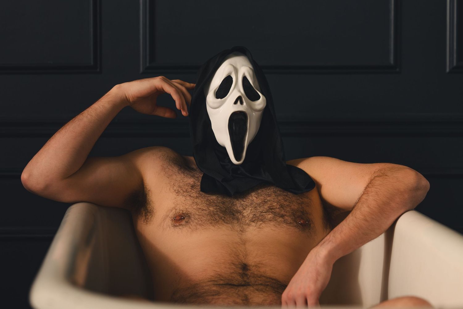 Person wearing a Ghostface Halloween mask sitting shirtless in a bathtub against a dark wall.