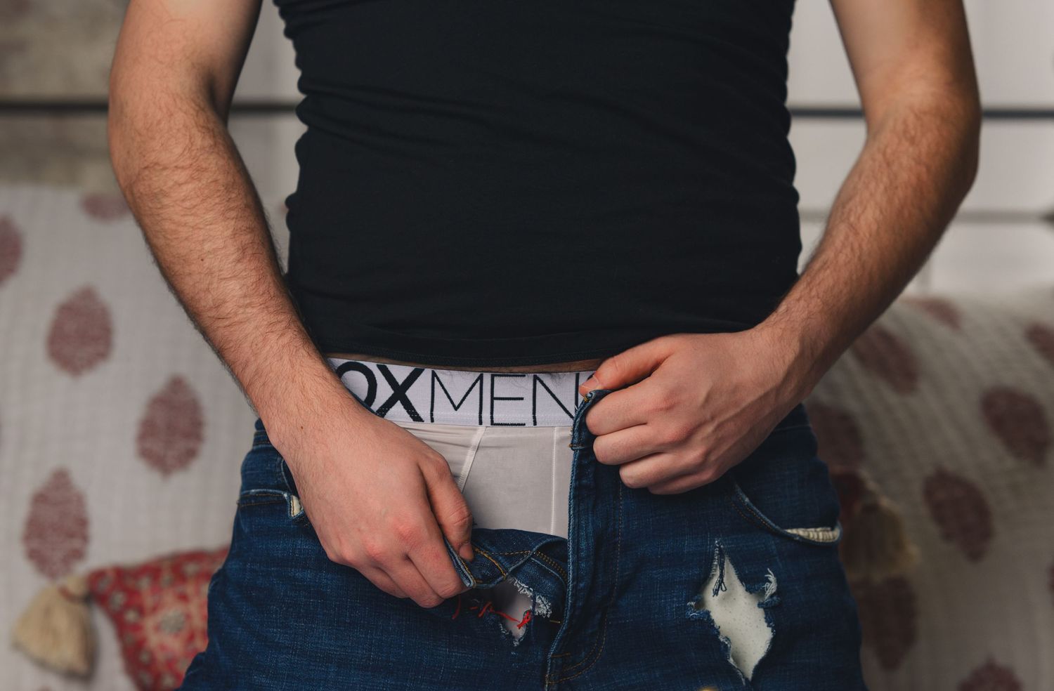 Man in black tank top and ripped jeans wearing branded underwear waistband visible above pants.