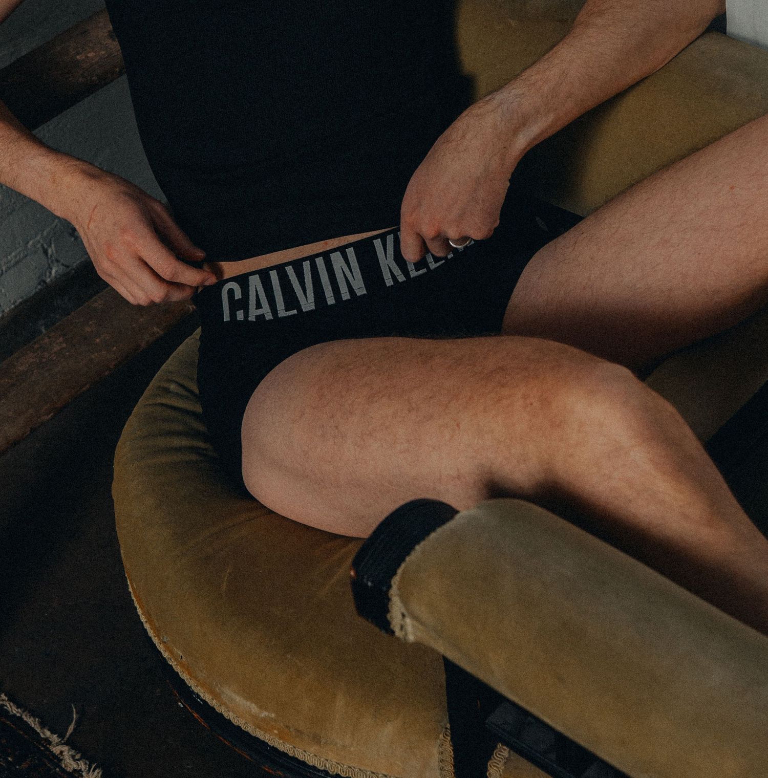 Man wearing Calvin Klein underwear while seated on a vintage olive green chair.
