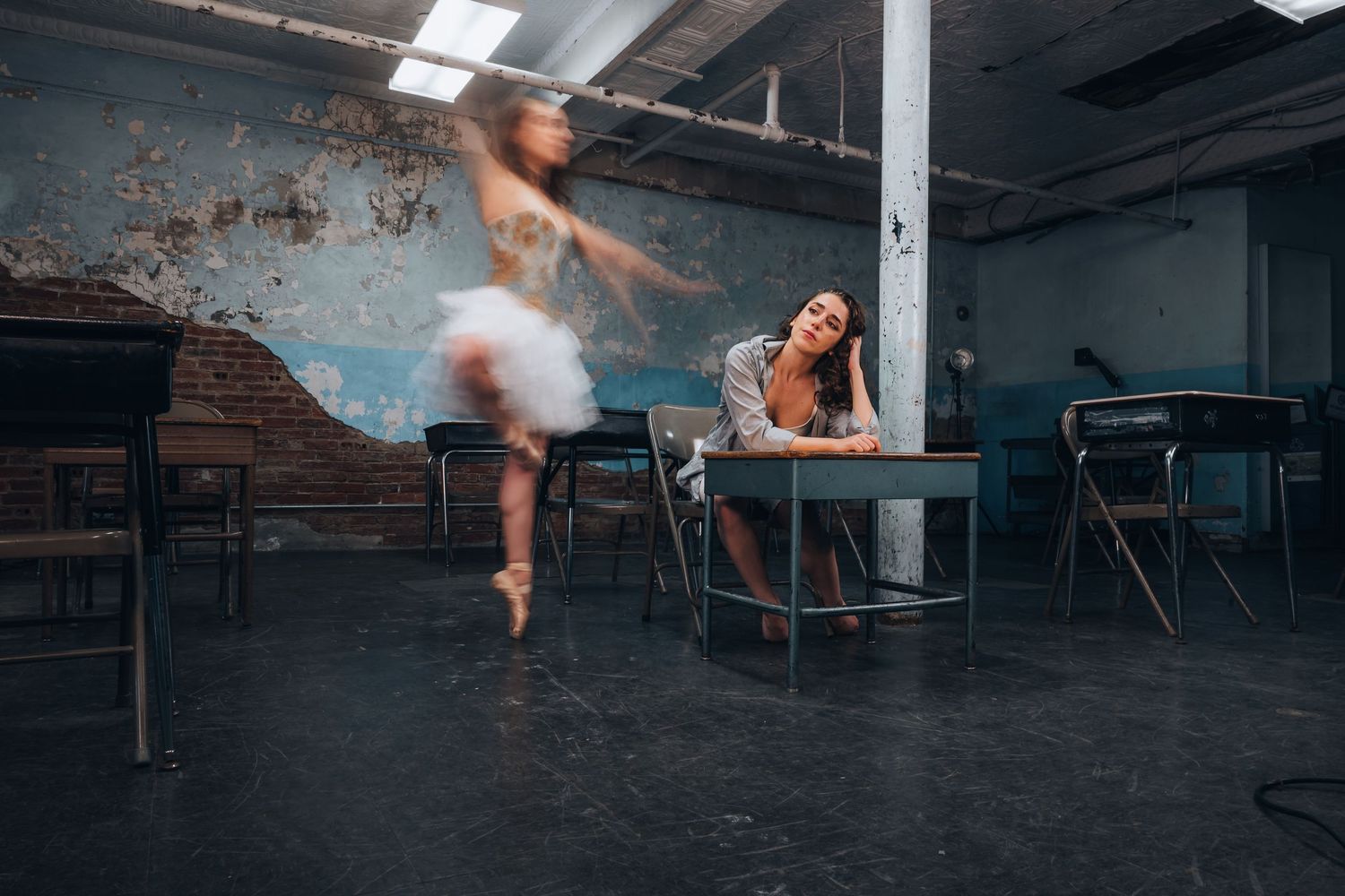 A ballerina in a white tutu spins gracefully while a woman rests at a desk in an urban studio.