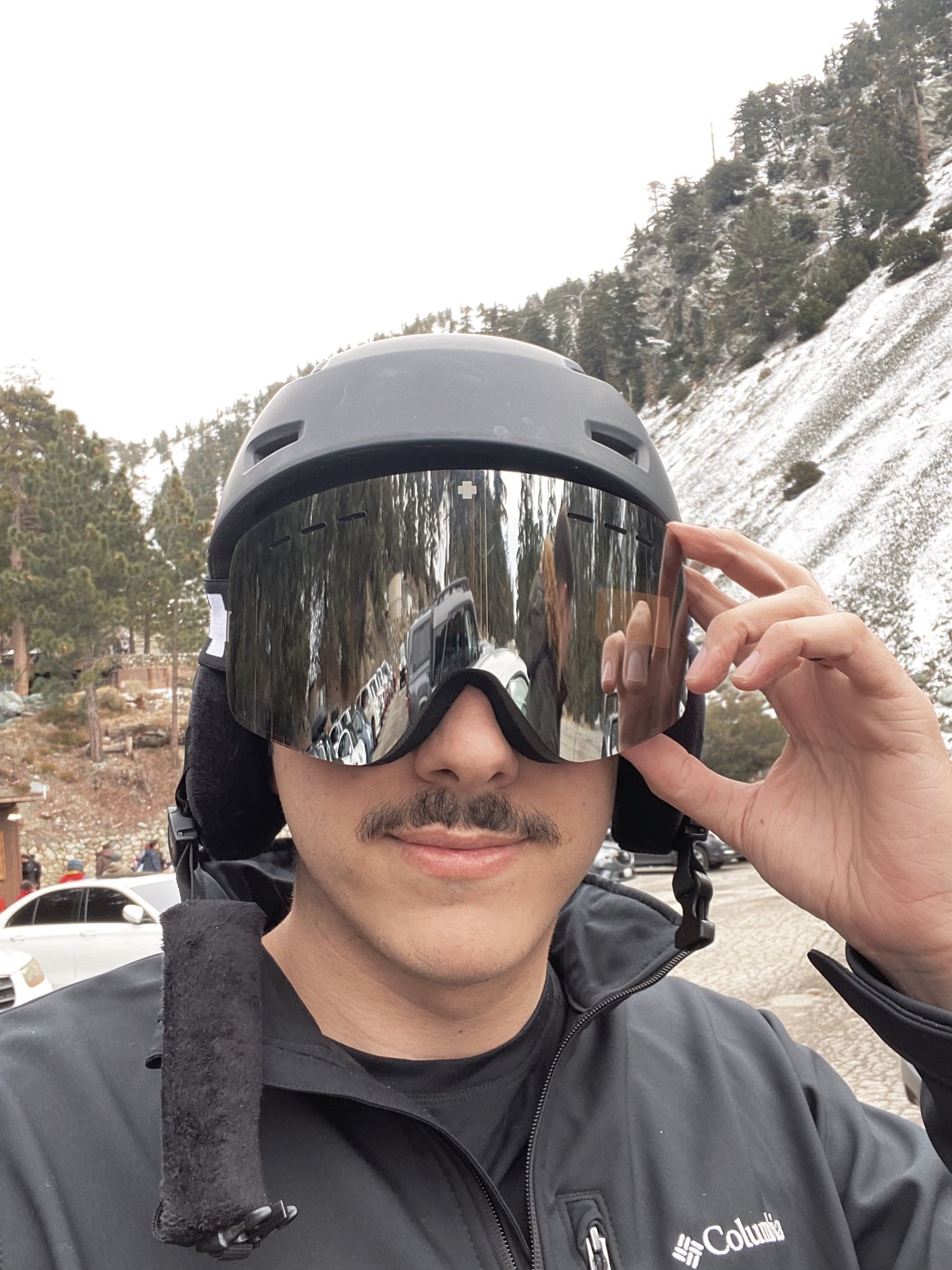 Man wearing black ski helmet and mirrored goggles with Columbia jacket on snowy mountain slope.