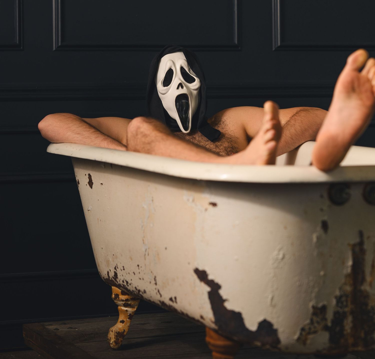 Person wearing a Ghostface Halloween mask relaxing in a vintage clawfoot bathtub in a dark room.