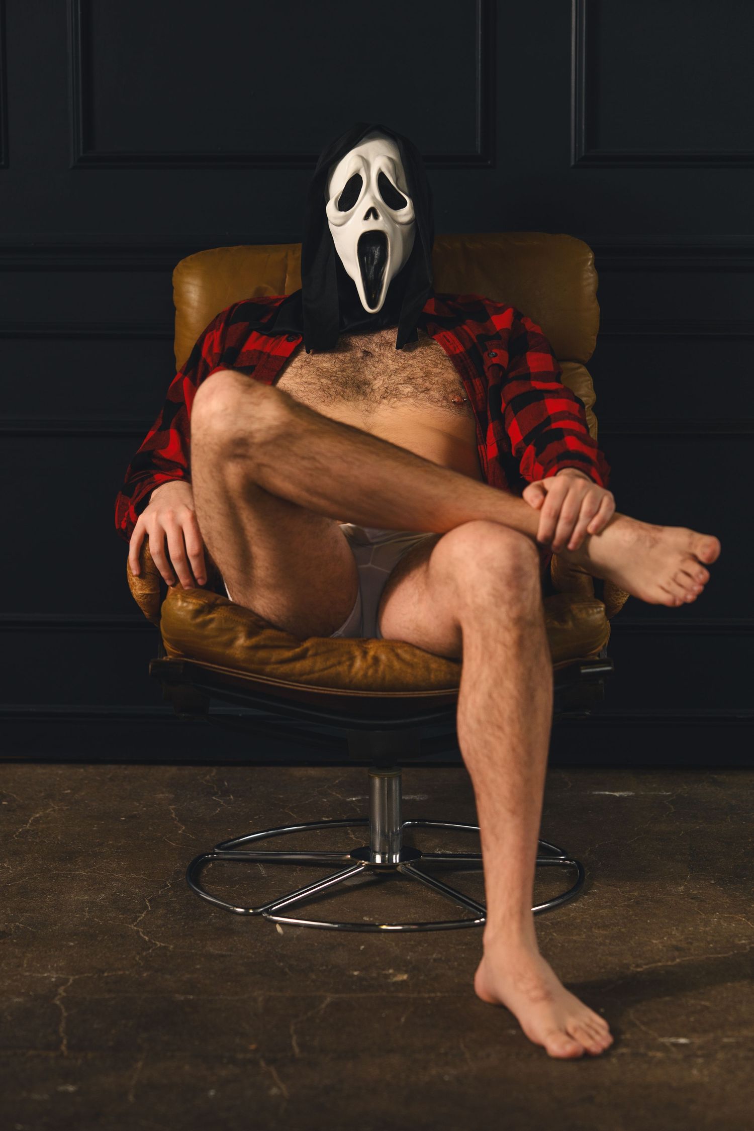 Person wearing a Ghostface Halloween mask and red flannel shirt sitting in a brown leather chair with legs crossed.
