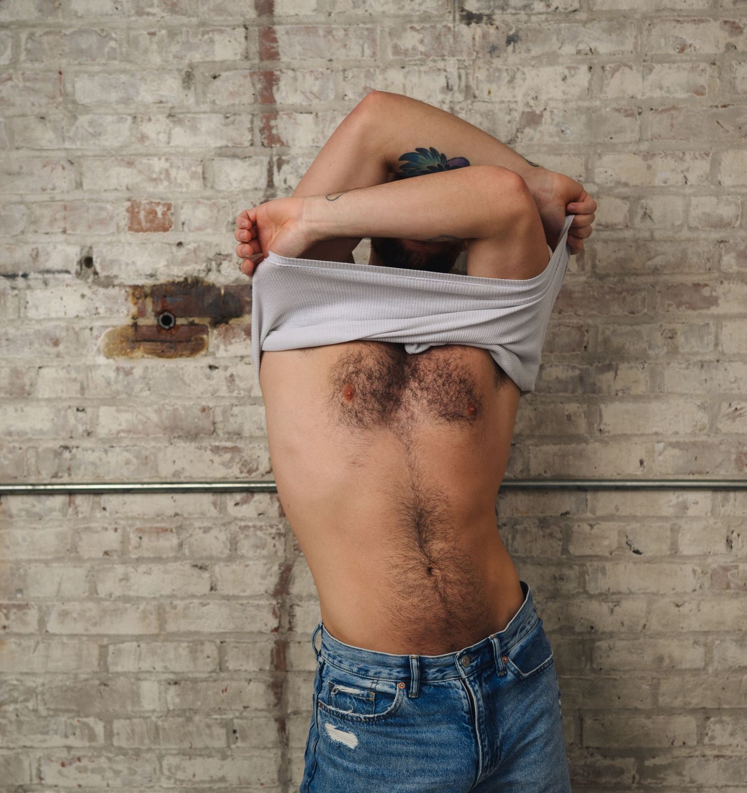 Person lifting white shirt to reveal torso with body hair, standing against rustic brick wall, wearing blue jeans.