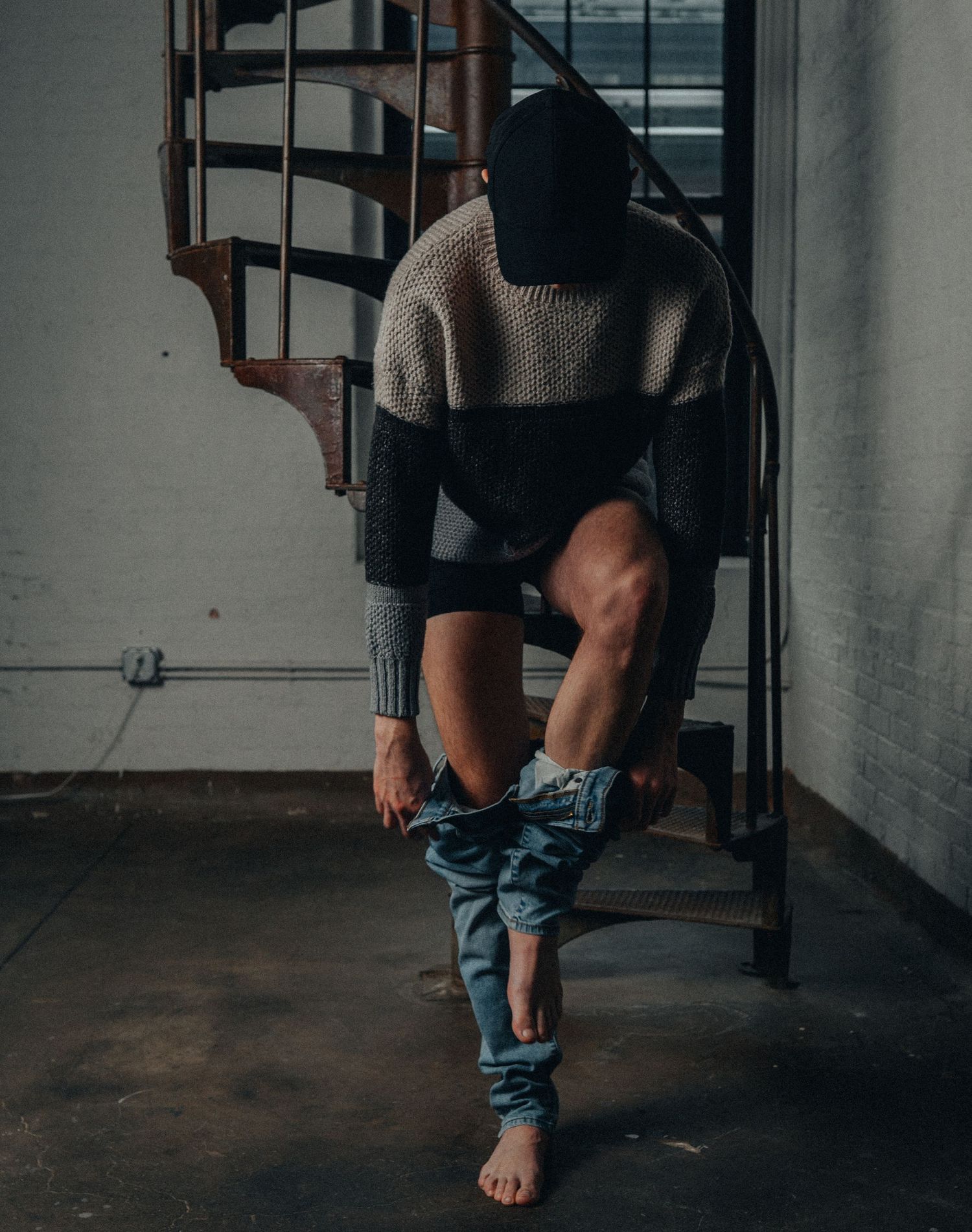 Person in knit sweater and black beanie sitting on steps of spiral staircase, pulling up jeans.