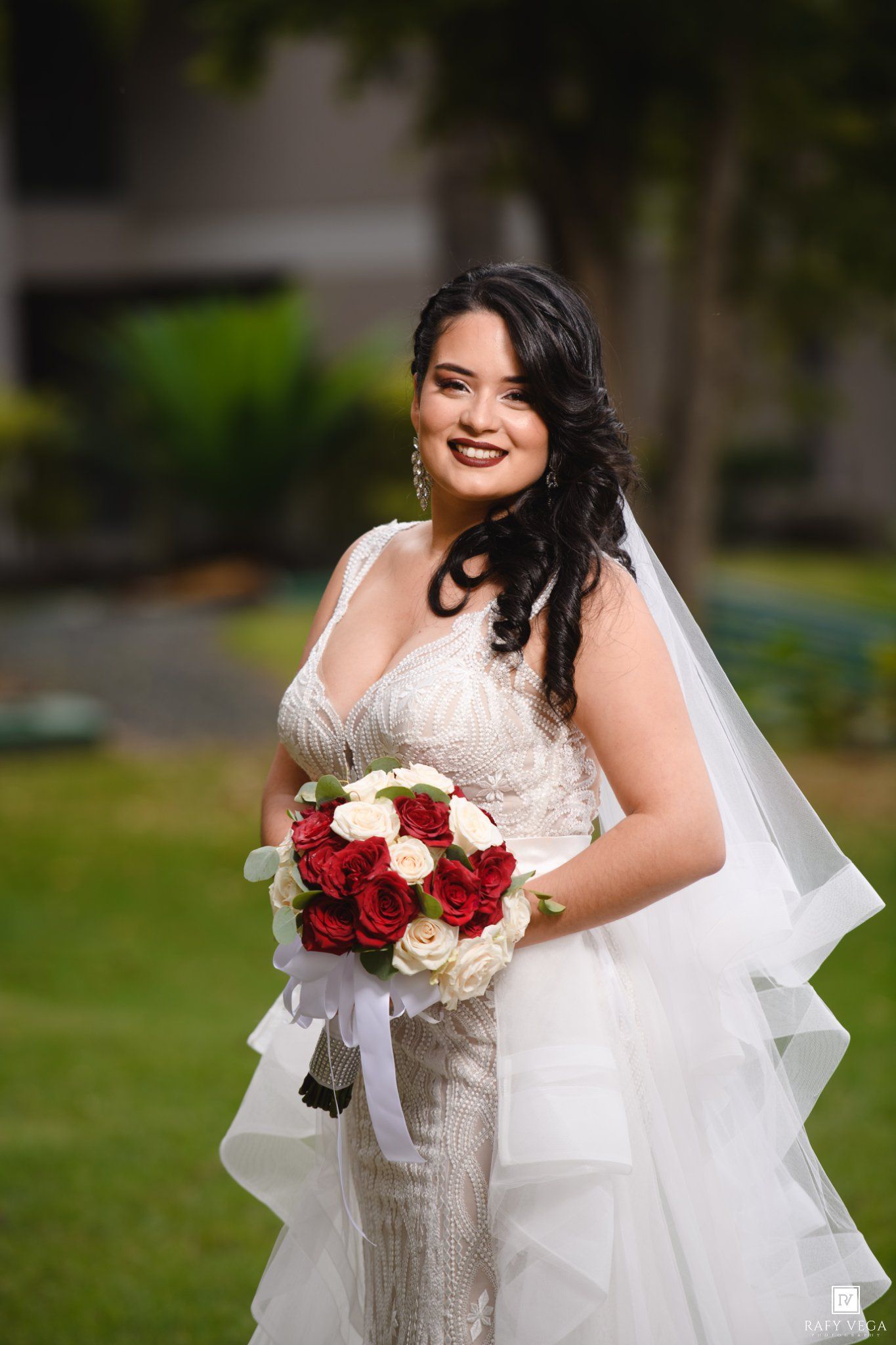 Bride in white lace gown holding red and white rose bouquet, smiling outdoors.