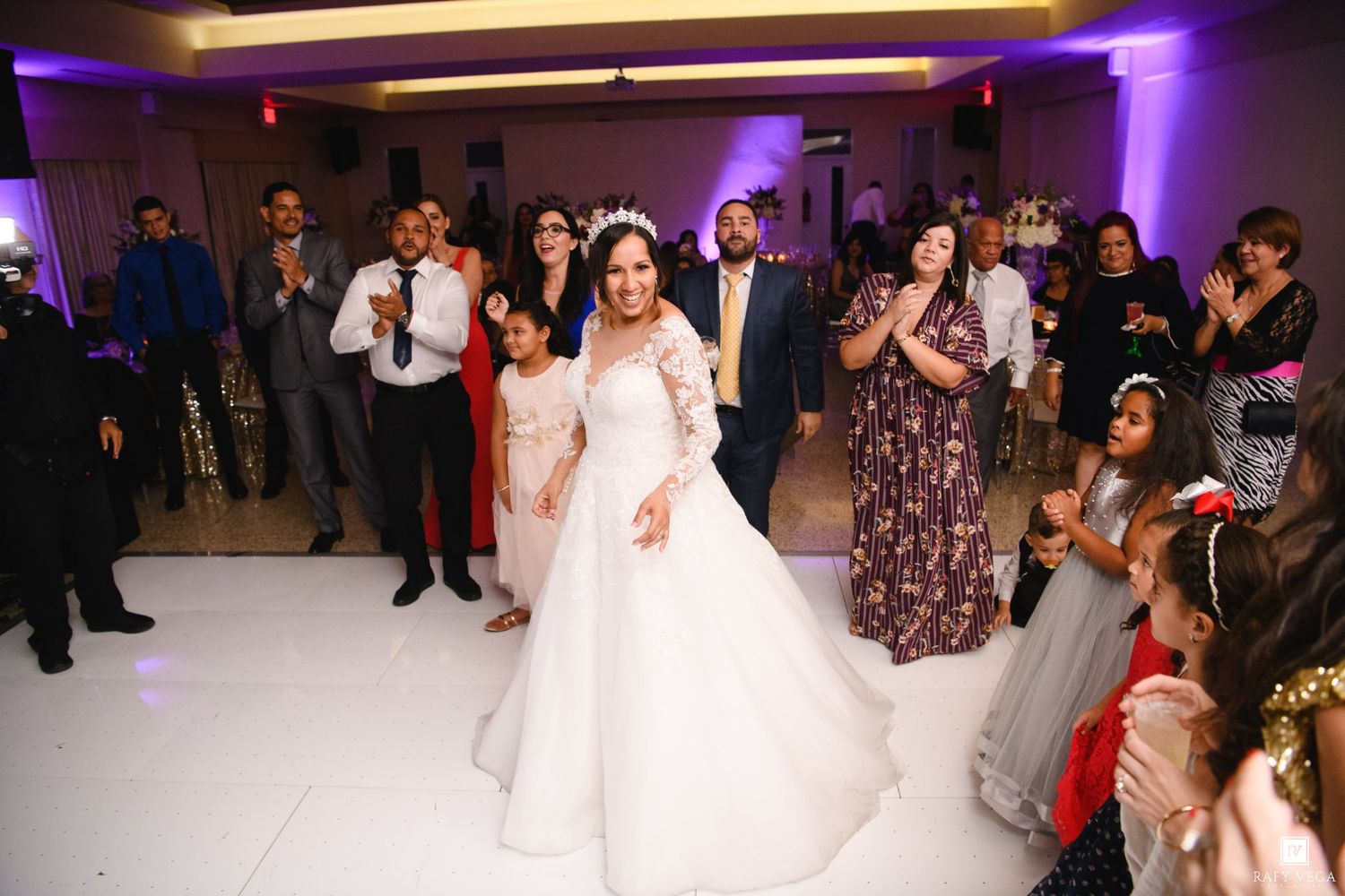 Bride in white ball gown dancing at wedding reception surrounded by smiling guests on purple-lit dance floor.