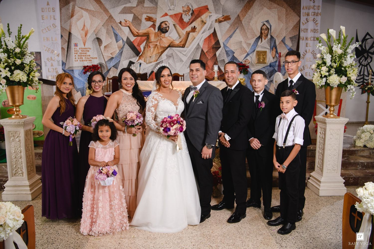 Wedding party poses in church, bride in white gown, bridesmaids in purple, groomsmen in suits, flower girl in pink.