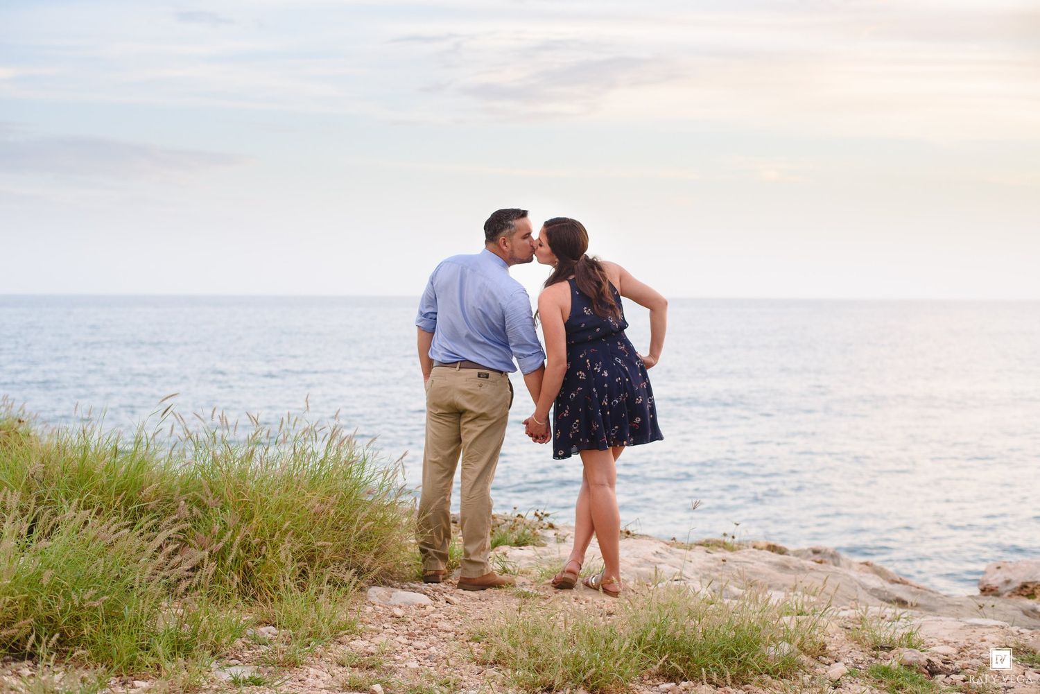 Couple kissing by the ocean shore at sunset near tall grass on rocky coastline.