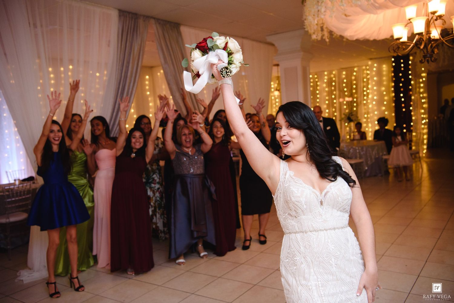 Bride tossing wedding bouquet to excited guests in elegant reception venue with fairy lights backdrop.