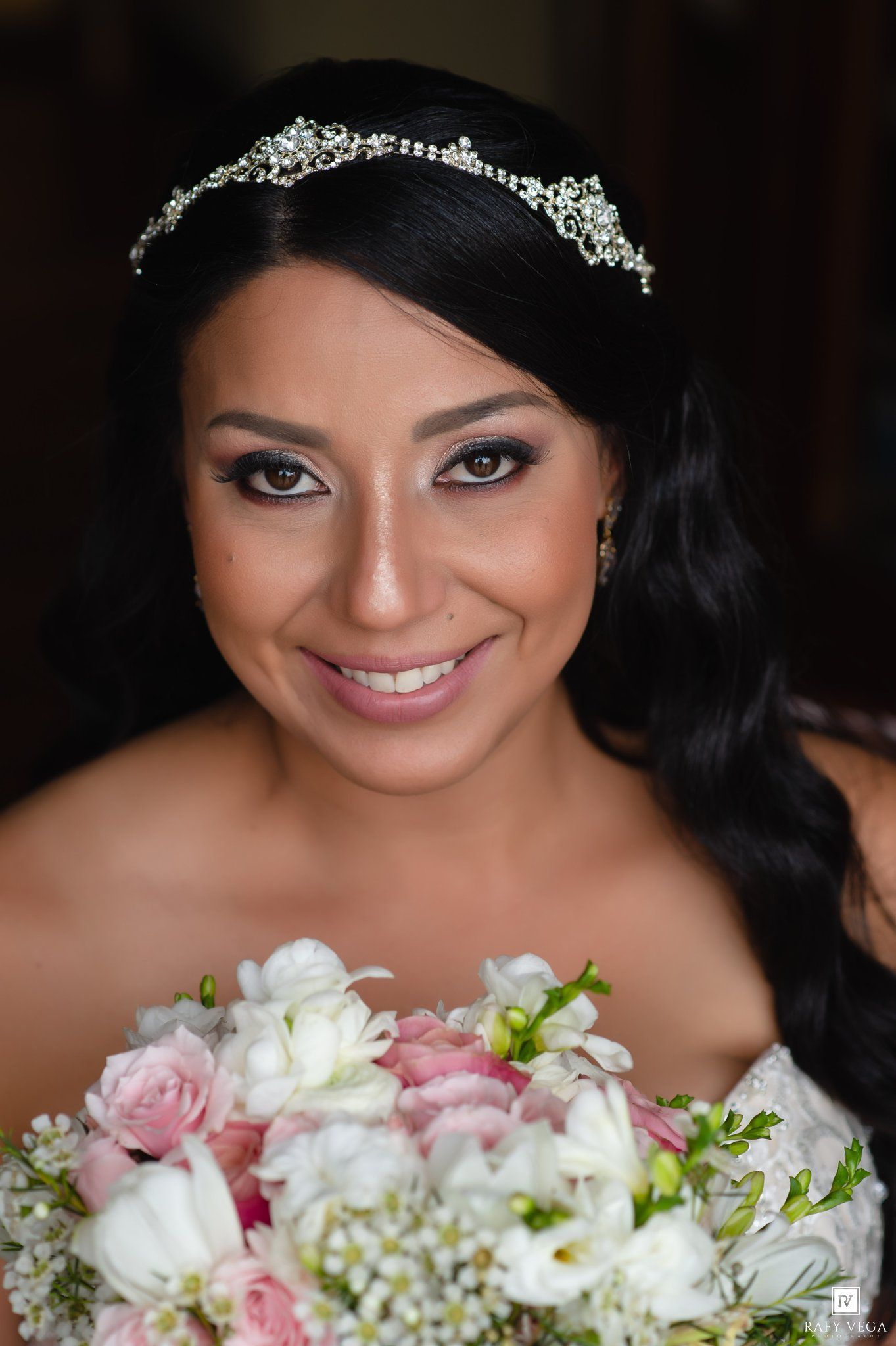 Smiling bride with crystal tiara and dark hair holding pink and white floral bouquet in elegant bridal portrait.
