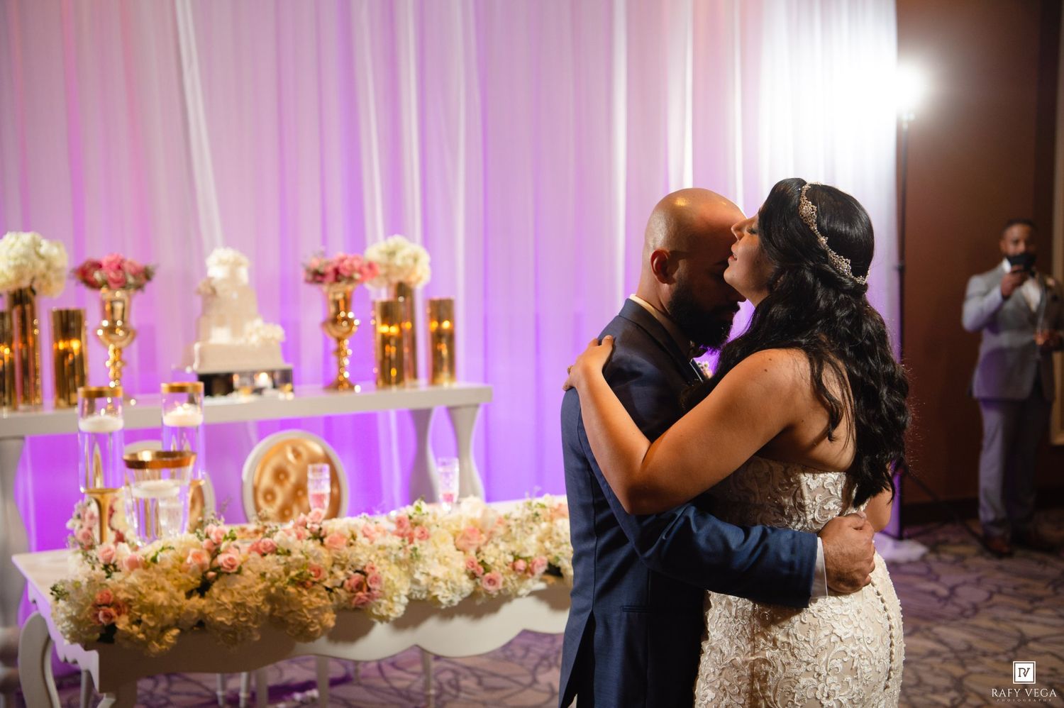 Bride and groom share a romantic first kiss at their elegant wedding reception with floral arrangements and gold decor.