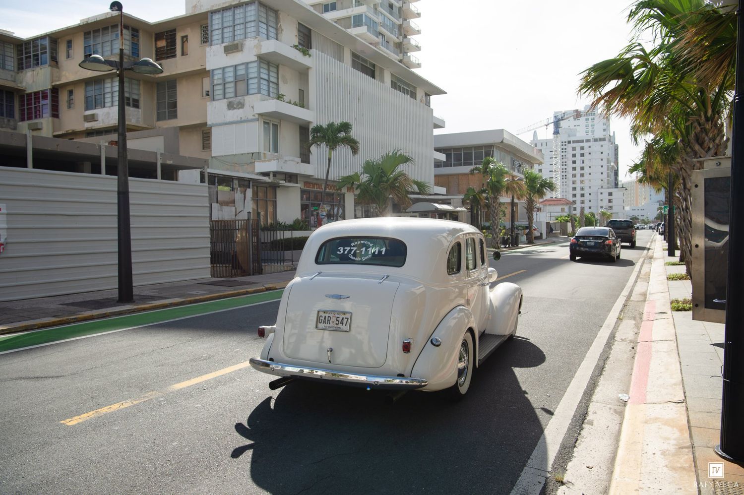 Vintage white classic car driving down a sunny urban street lined with palm trees and modern buildings.