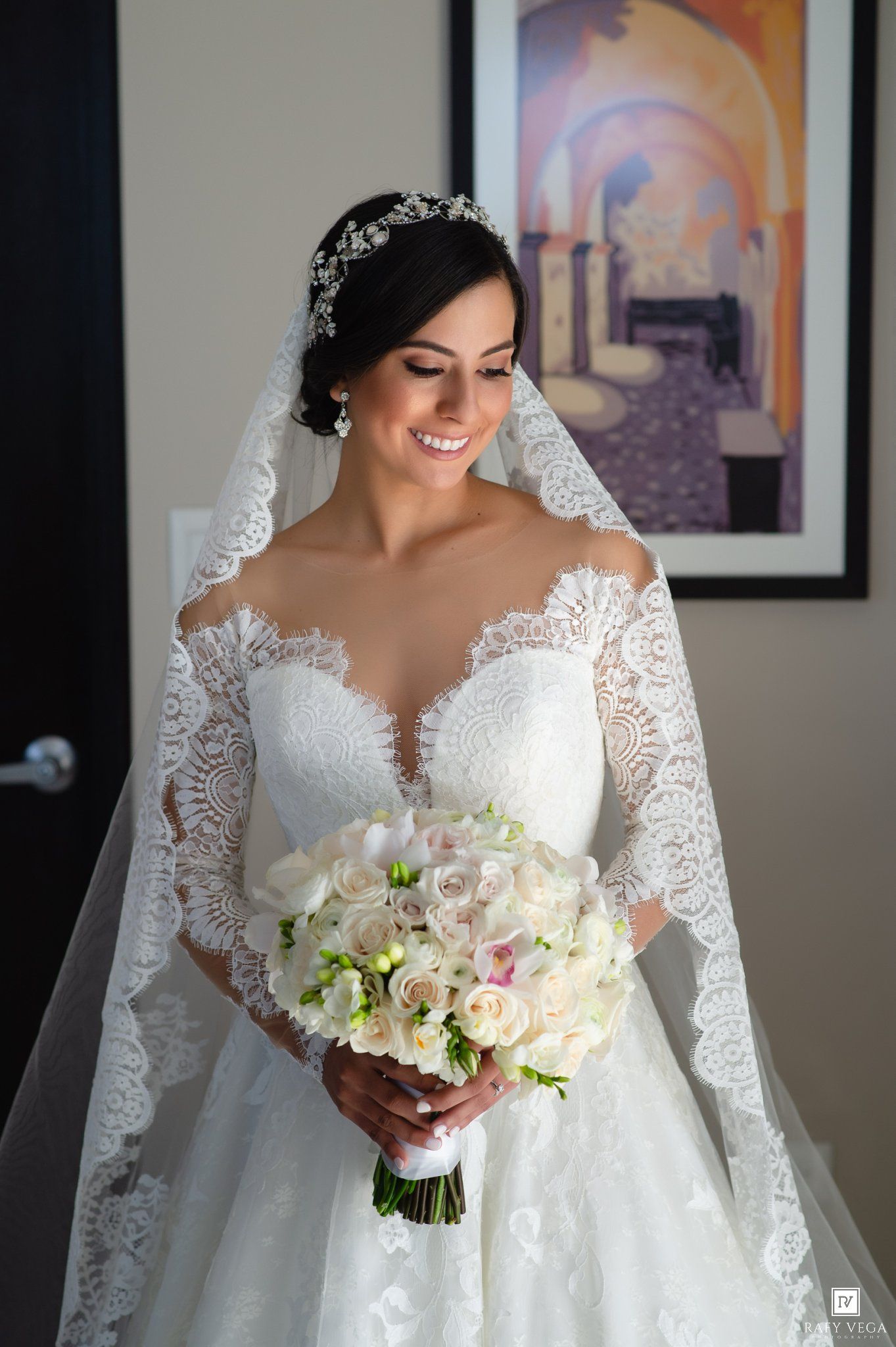 Bride in lace long-sleeve wedding gown with veil and tiara holding white floral bouquet, smiling elegantly indoors.