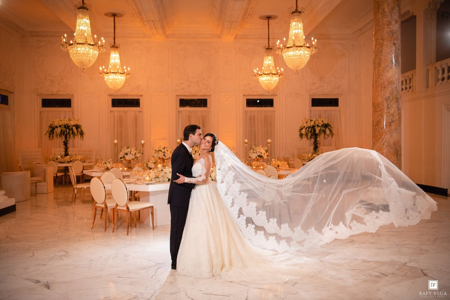 Bride and groom share romantic embrace in elegant ballroom, her lace veil flowing dramatically behind them.