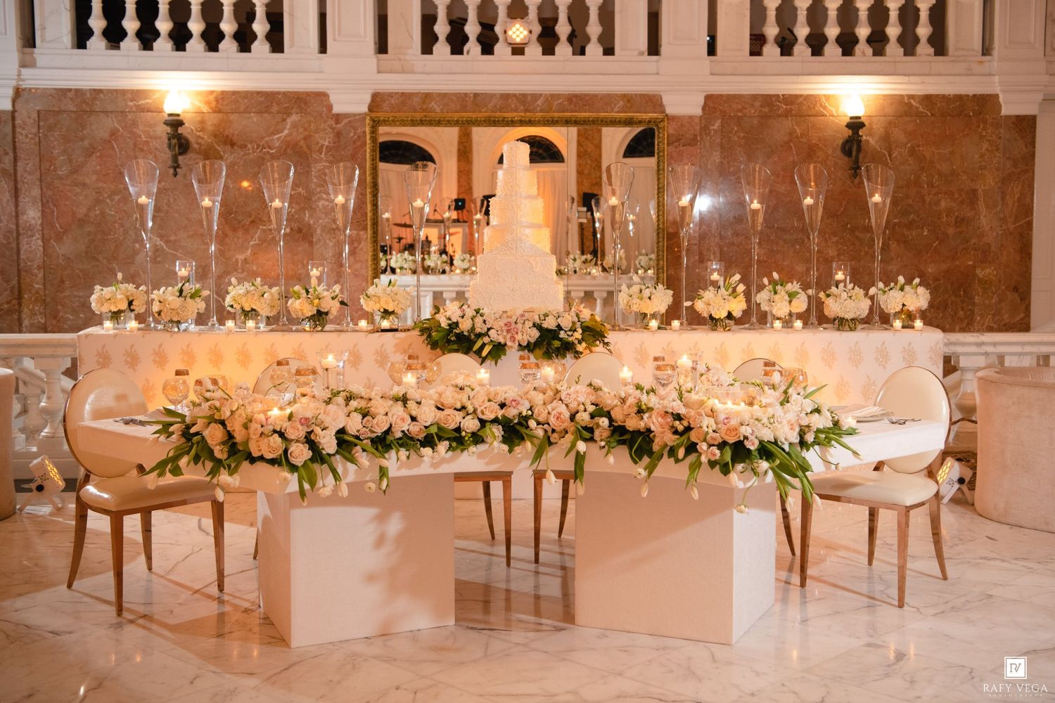 Elegant wedding head table adorned with lush blush floral arrangements, tall crystal vases, and a white tiered cake in a grand ballroom.
