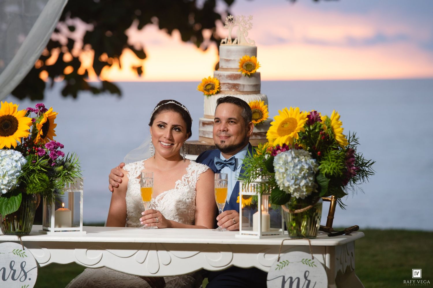 Bride and groom sit at sweetheart table with sunflowers, wedding cake, and ocean sunset backdrop.