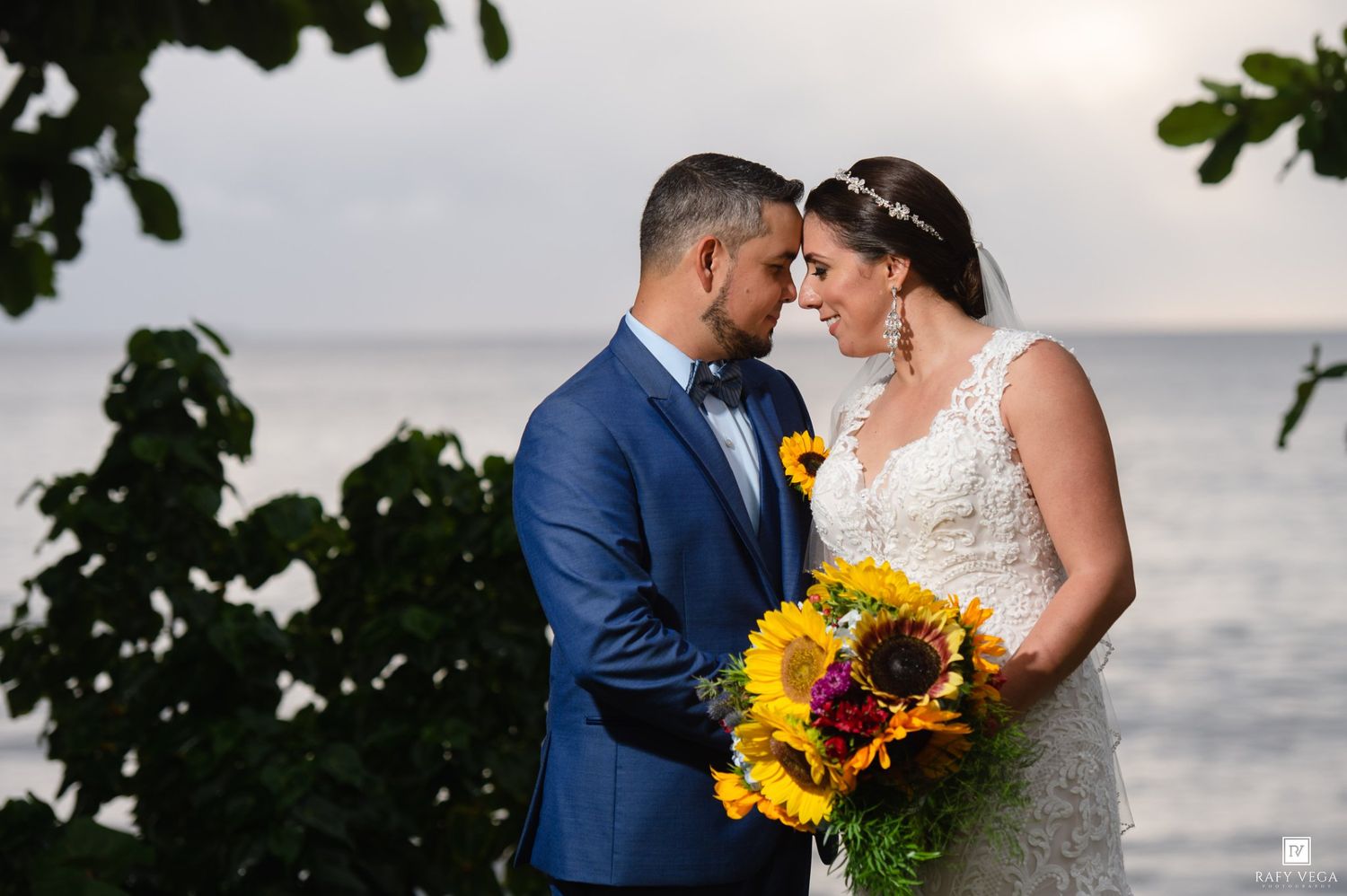 Bride and groom share a romantic moment by the ocean, she holds a vibrant sunflower bouquet, he wears a blue suit.
