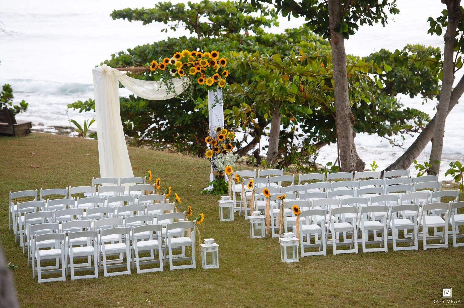 Outdoor beach wedding ceremony setup with white chairs, lanterns, and a floral arch decorated with sunflowers by the ocean.
