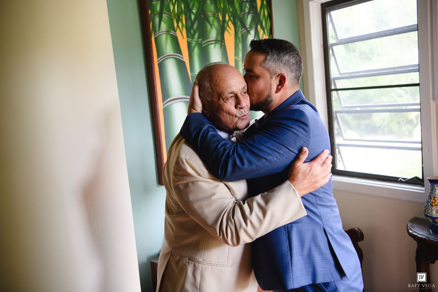 Two men in suits share a warm embrace indoors, one kissing the other on the cheek in an emotional moment.