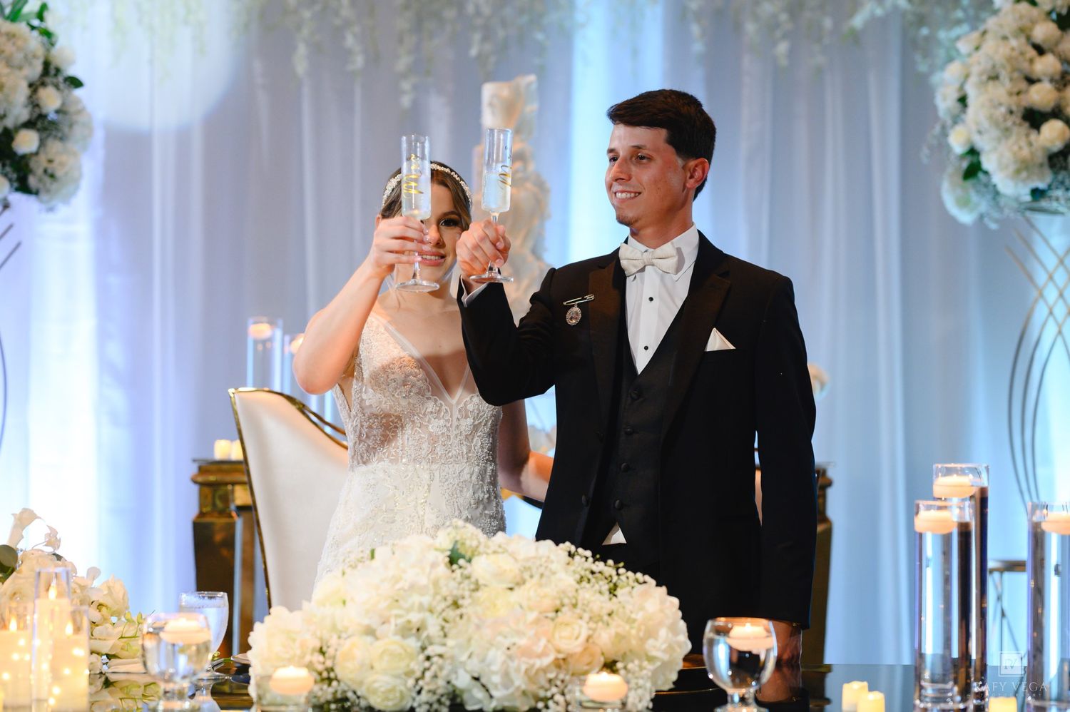 Bride and groom toasting with champagne glasses at elegant white floral wedding reception.