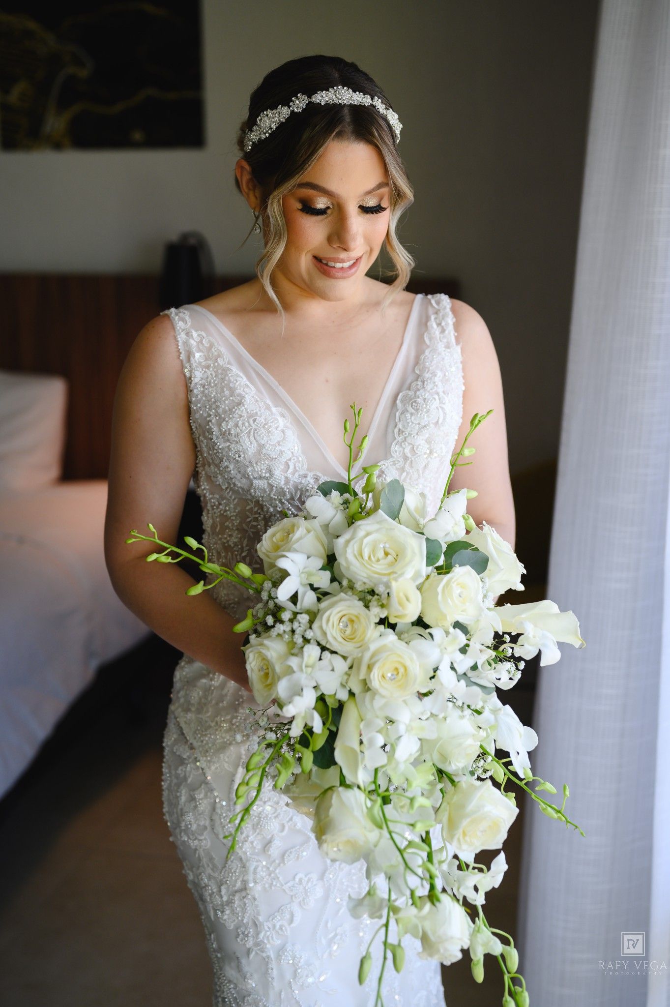 Bride in beaded gown holding cascading white rose bouquet with greenery, wearing crystal headpiece on wedding day.