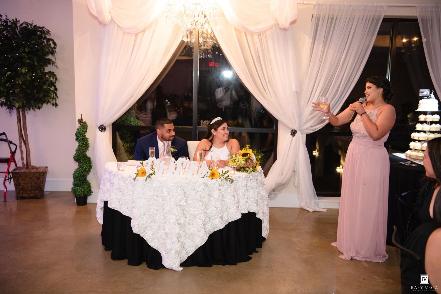 Bride and groom seated at sweetheart table while bridesmaid in pink gives a toast at elegant wedding reception.