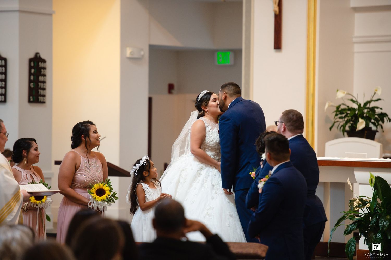 Bride and groom share a kiss at the altar during a church wedding ceremony with bridesmaids and flower girls nearby.