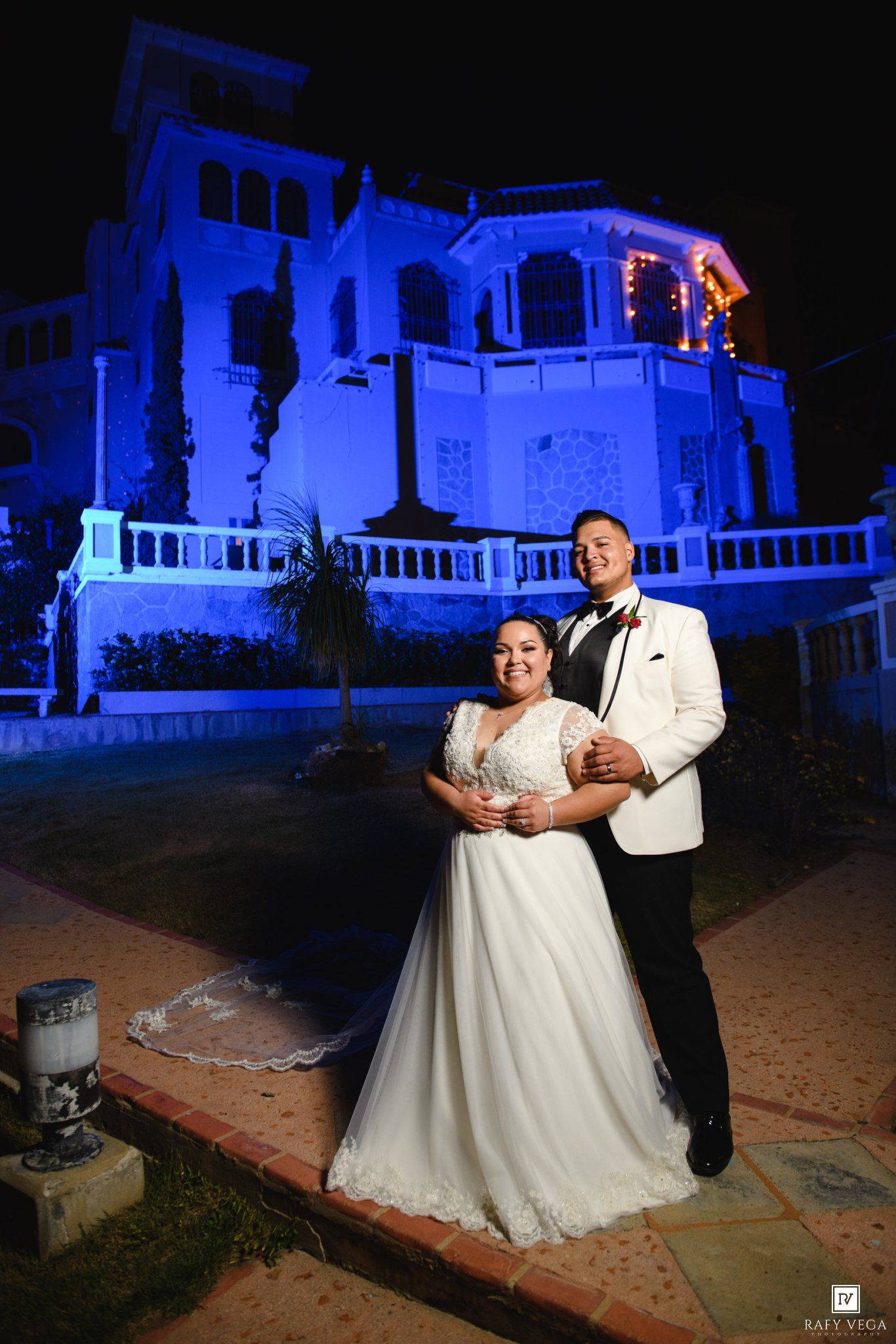 Bride in white gown and groom in white tuxedo pose at night in front of a blue-lit mansion venue.