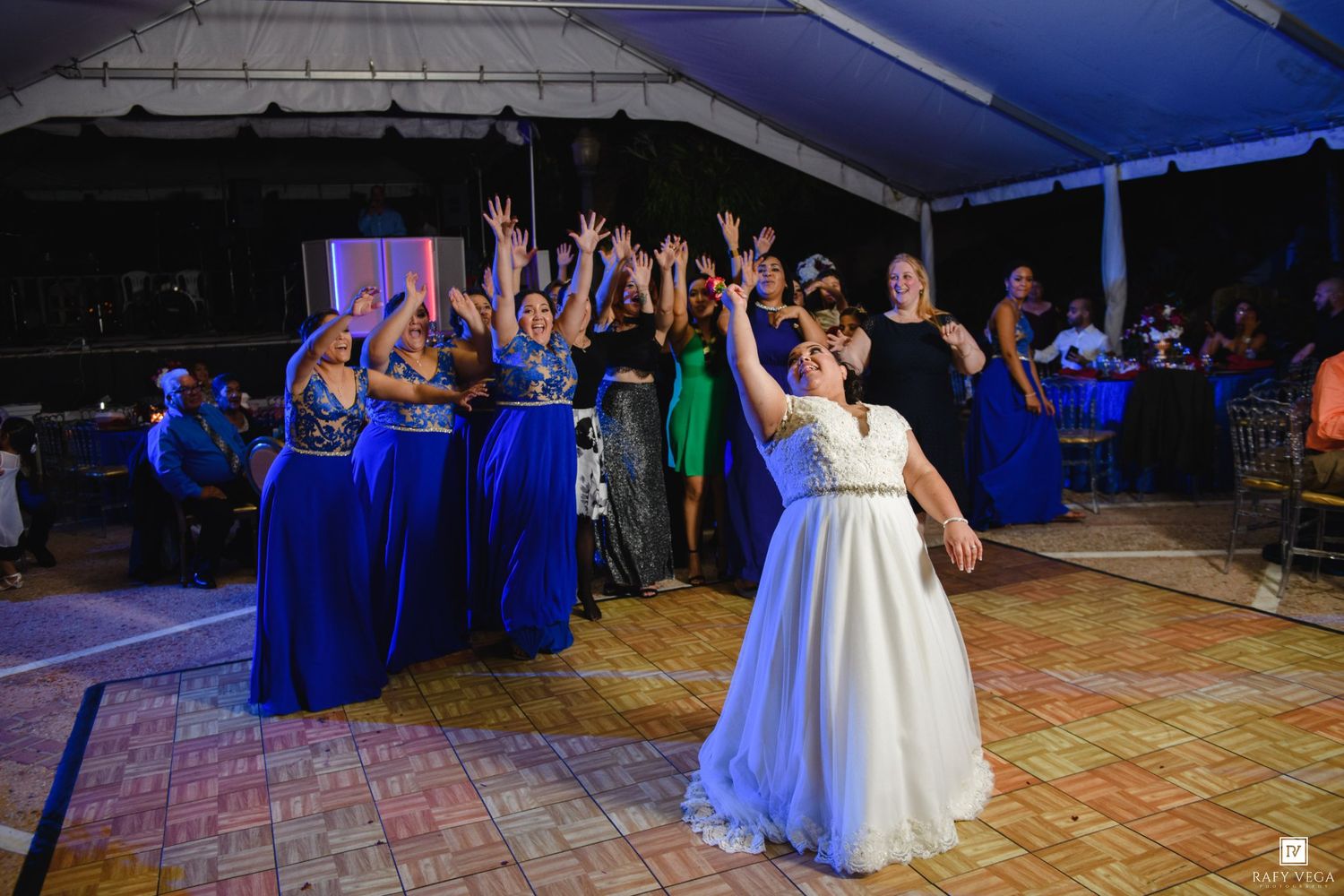 Bride tossing bouquet to excited bridesmaids in blue dresses at an outdoor wedding reception.
