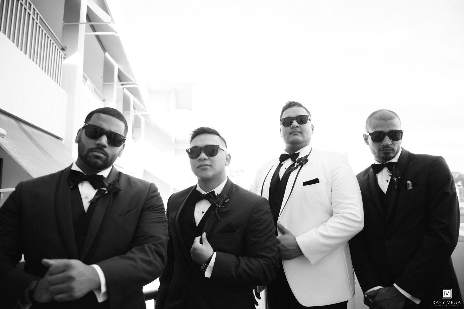 Four groomsmen in black and white tuxedos wearing sunglasses pose outdoors.