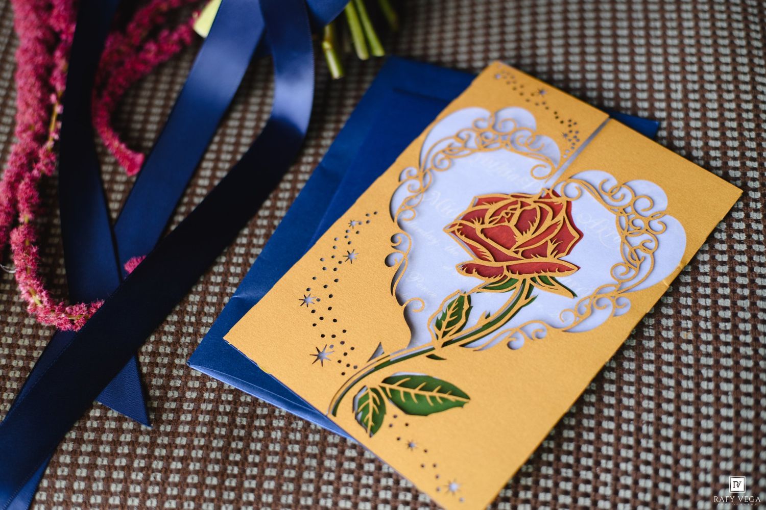 Elegant gold wedding card with a red rose design and blue ribbon on a decorative surface.