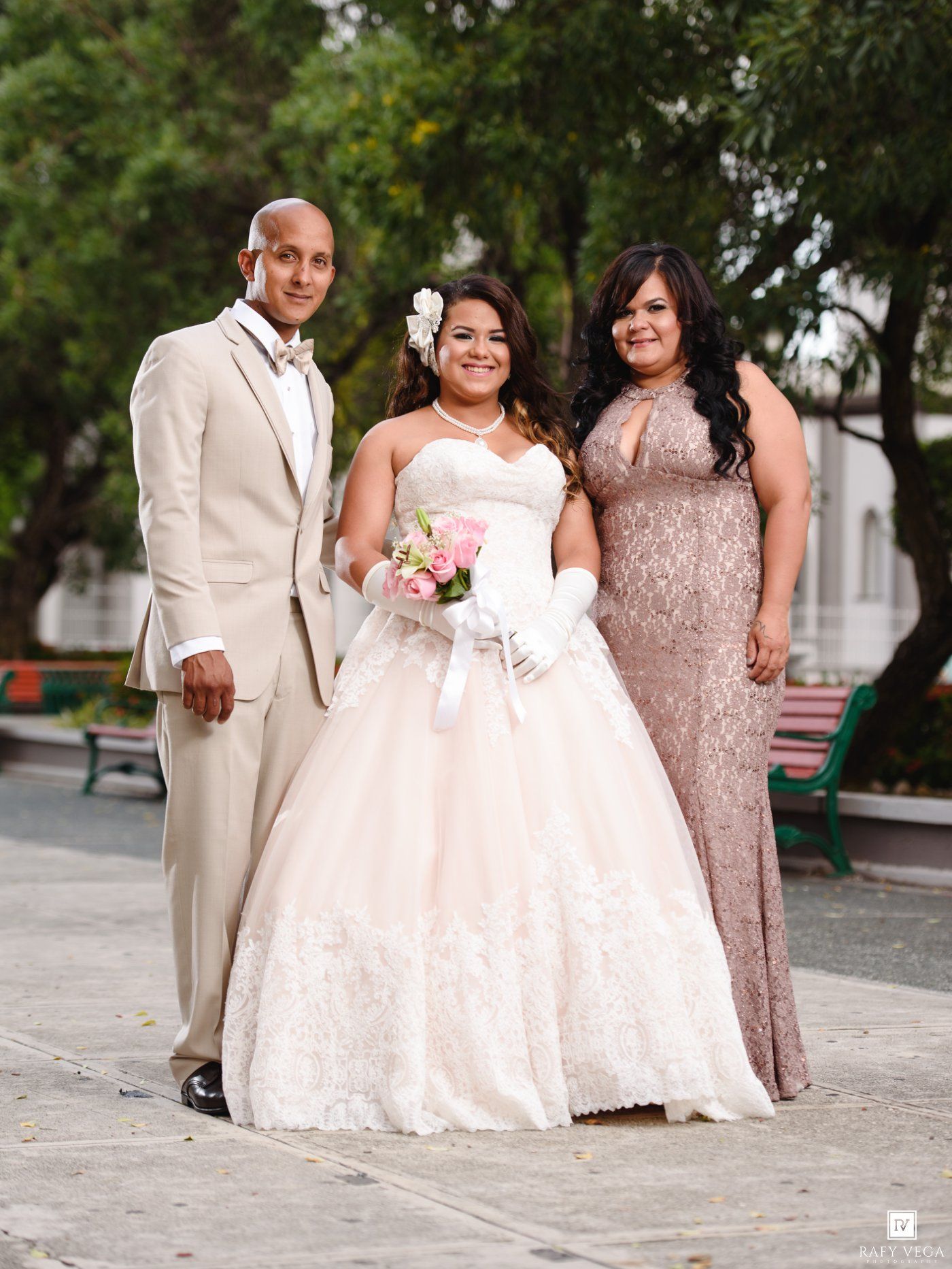 Young woman in white quinceañera gown holding pink bouquet poses with formally dressed man and woman outdoors.