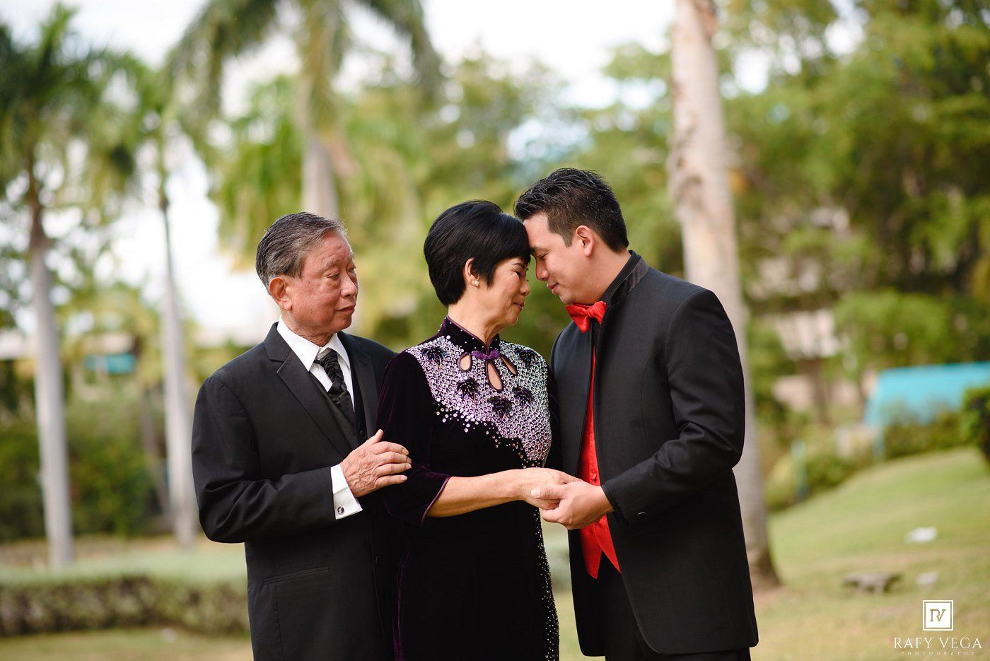 Three formally dressed people converse outdoors near palm trees, with two men in suits and a woman in a floral dress.