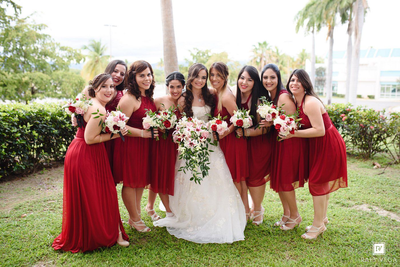 Bride with seven bridesmaids in red dresses holding bouquets posing outdoors on green lawn.