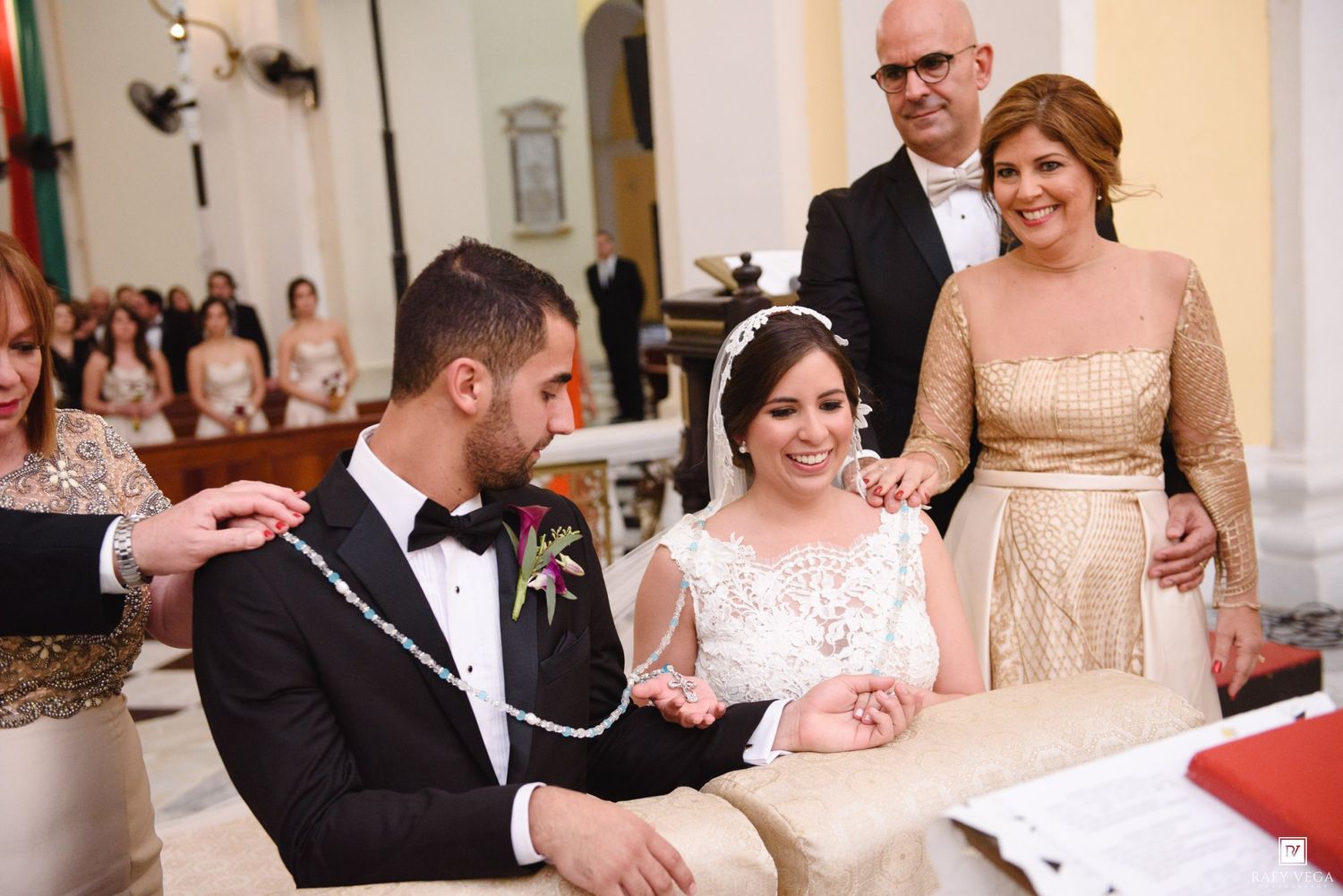 Bride and groom exchanging wedding ceremony unity chain at elegant church altar with family members looking on.