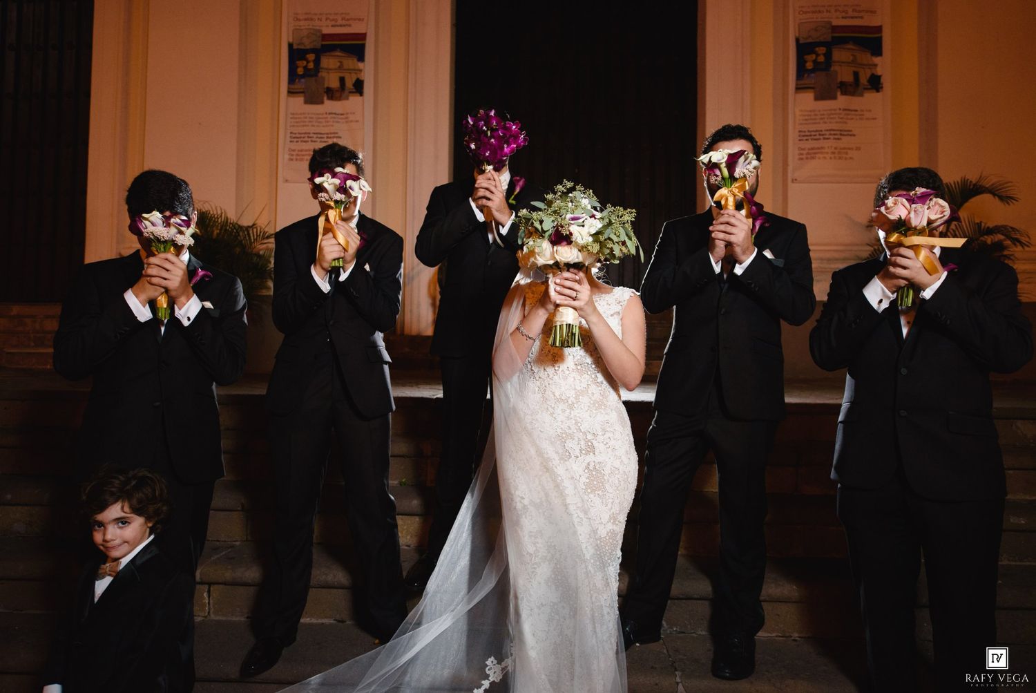 Bride holding purple bouquet surrounded by groomsmen in black suits wearing masquerade masks on church steps.