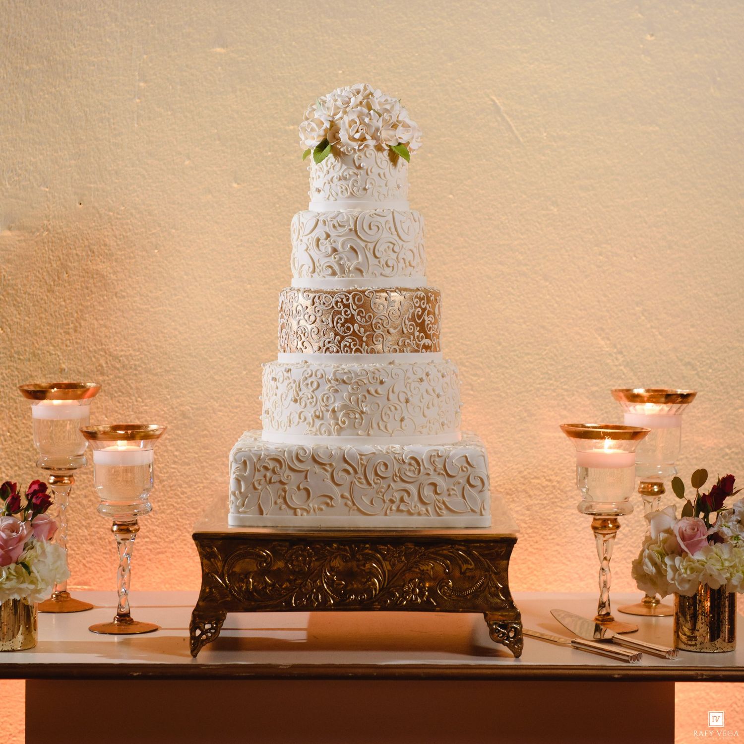 Elegant five-tier white and gold wedding cake with floral topper displayed on ornate stand surrounded by candles.