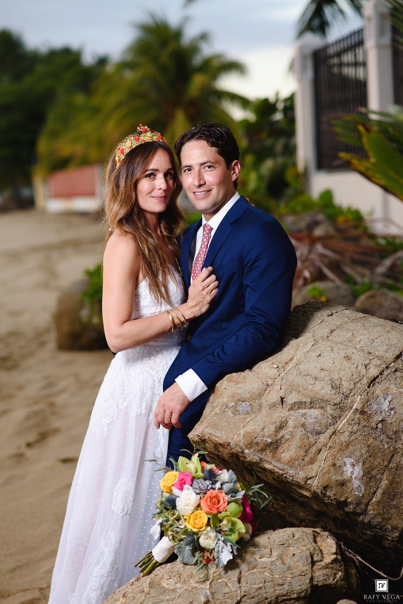 Bride and groom posing on a sandy beach with tropical palms, she holds a colorful bouquet, both dressed elegantly.