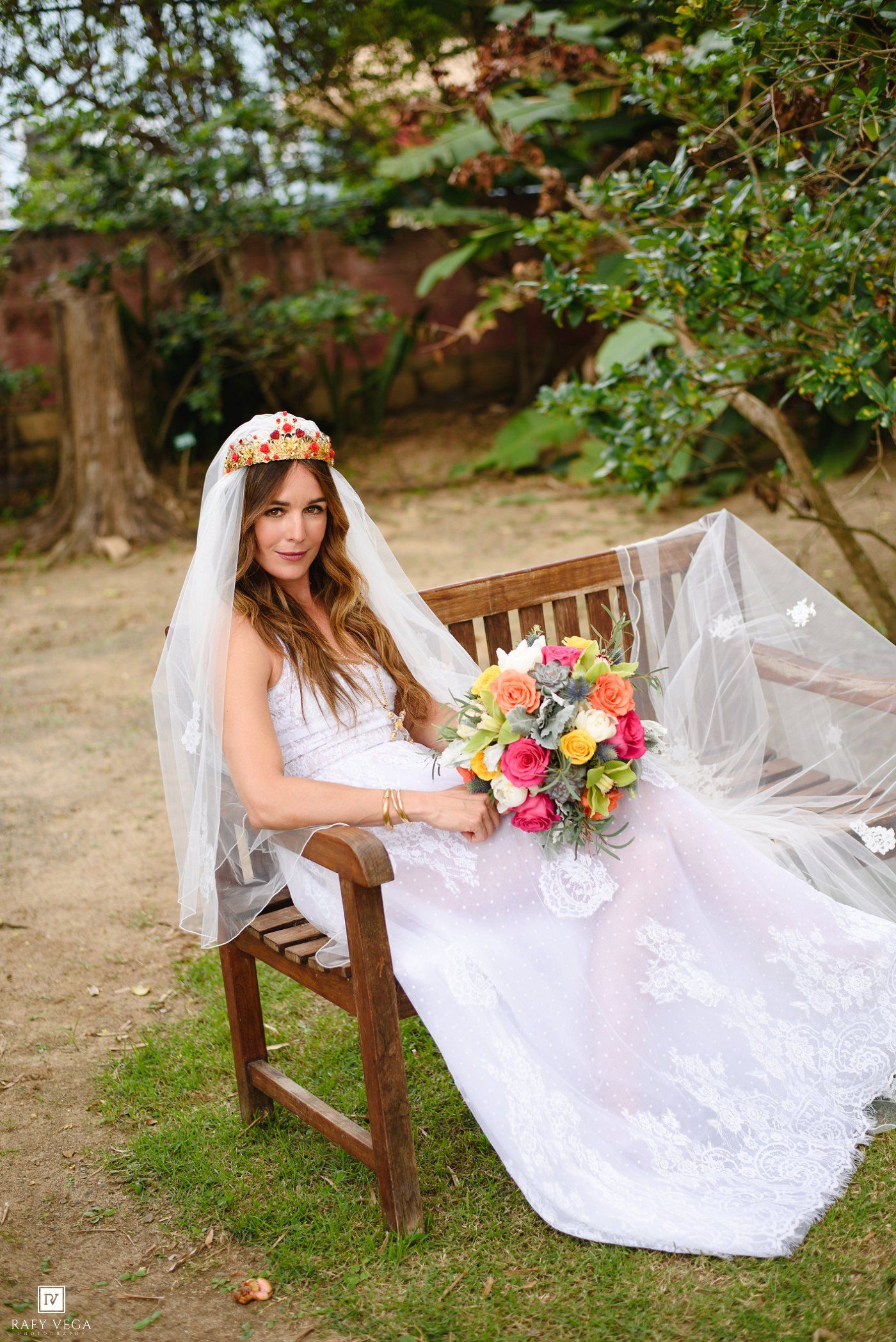 Bride in white lace gown and floral crown sits on wooden bench holding colorful bouquet outdoors.
