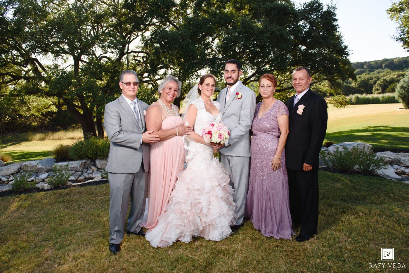 Wedding party posing outdoors on lush green lawn with large oak trees, bride in white gown with pink bouquet.